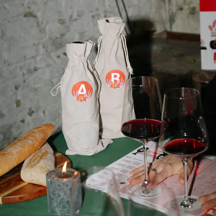 Twee anonieme flessen wijn in stoffen zakjes met de letters A en B, naast twee glazen rode wijn