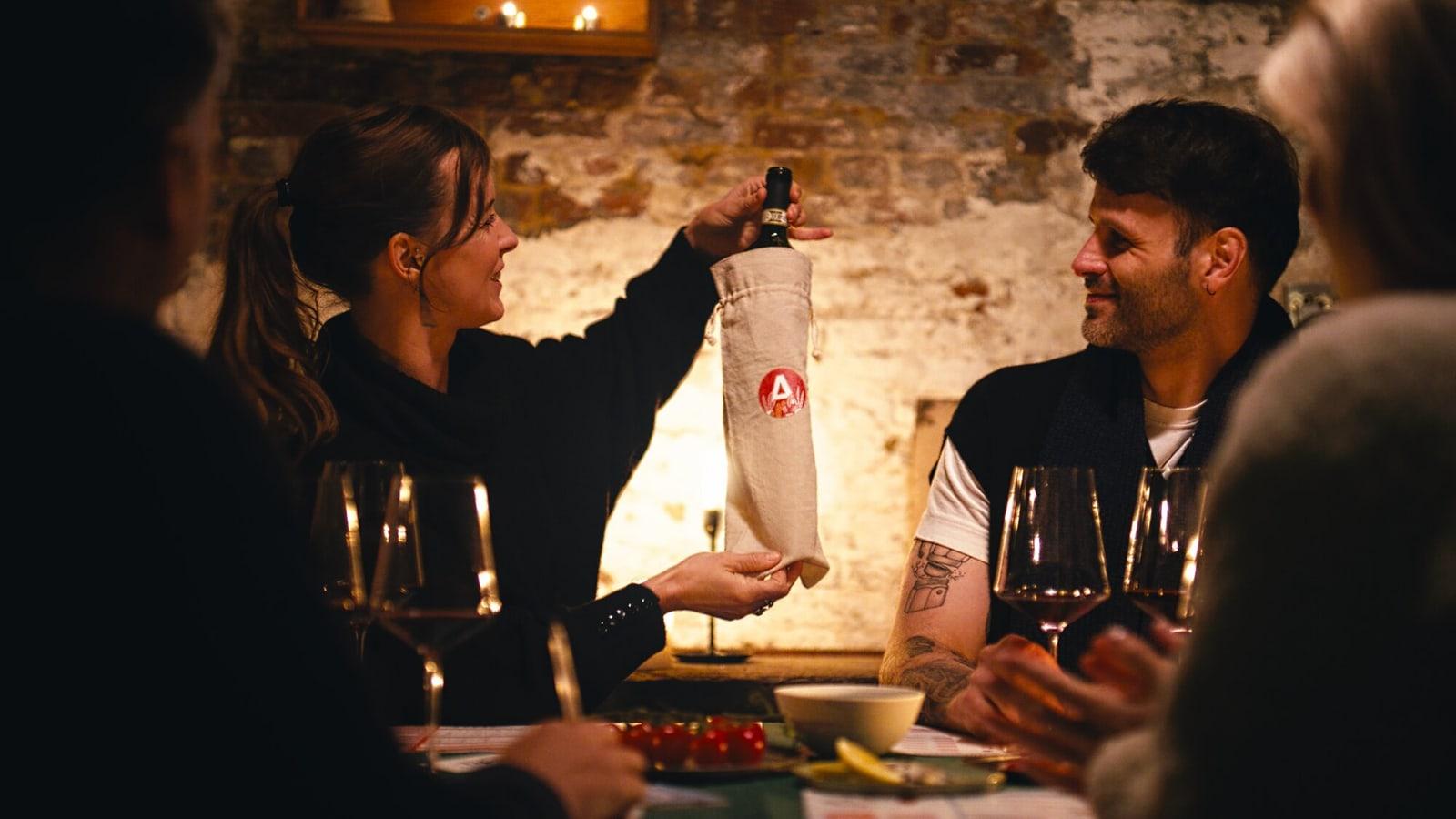 Vrienden aan tafel terwijl iemand een fles in een stoffen zakje omhooghoudt