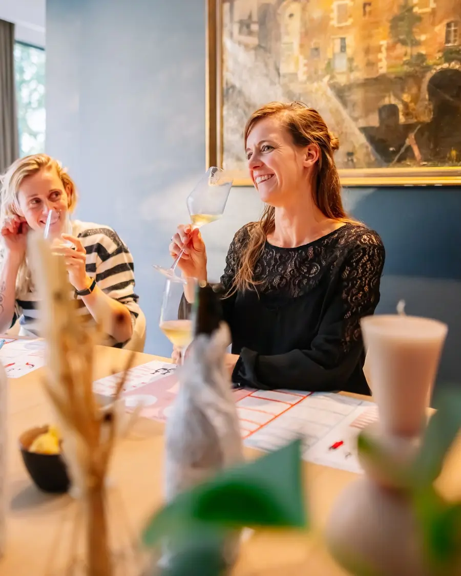 Twee vrouwen proeven witte wijn aan tafel, met een fles in een stoffen zakje ervoor