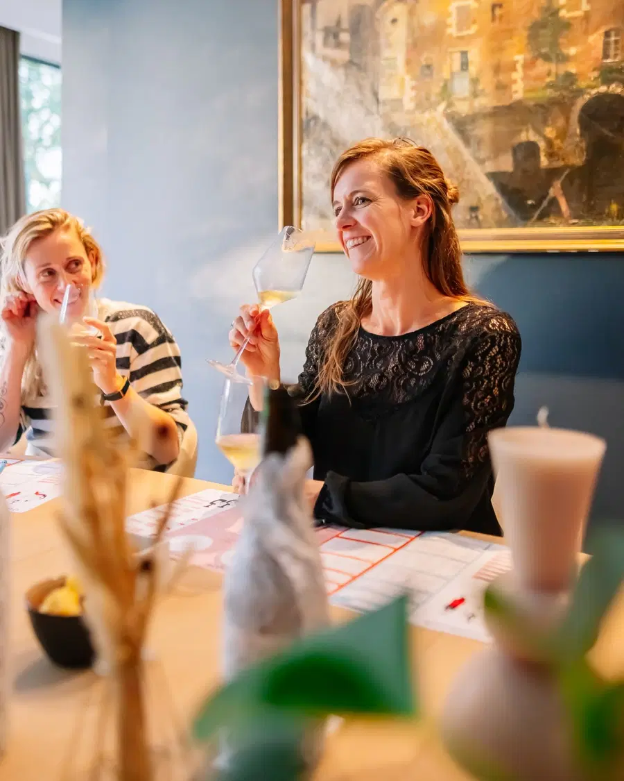 Twee vrouwen proeven witte wijn aan tafel, met een fles in een stoffen zakje ervoor